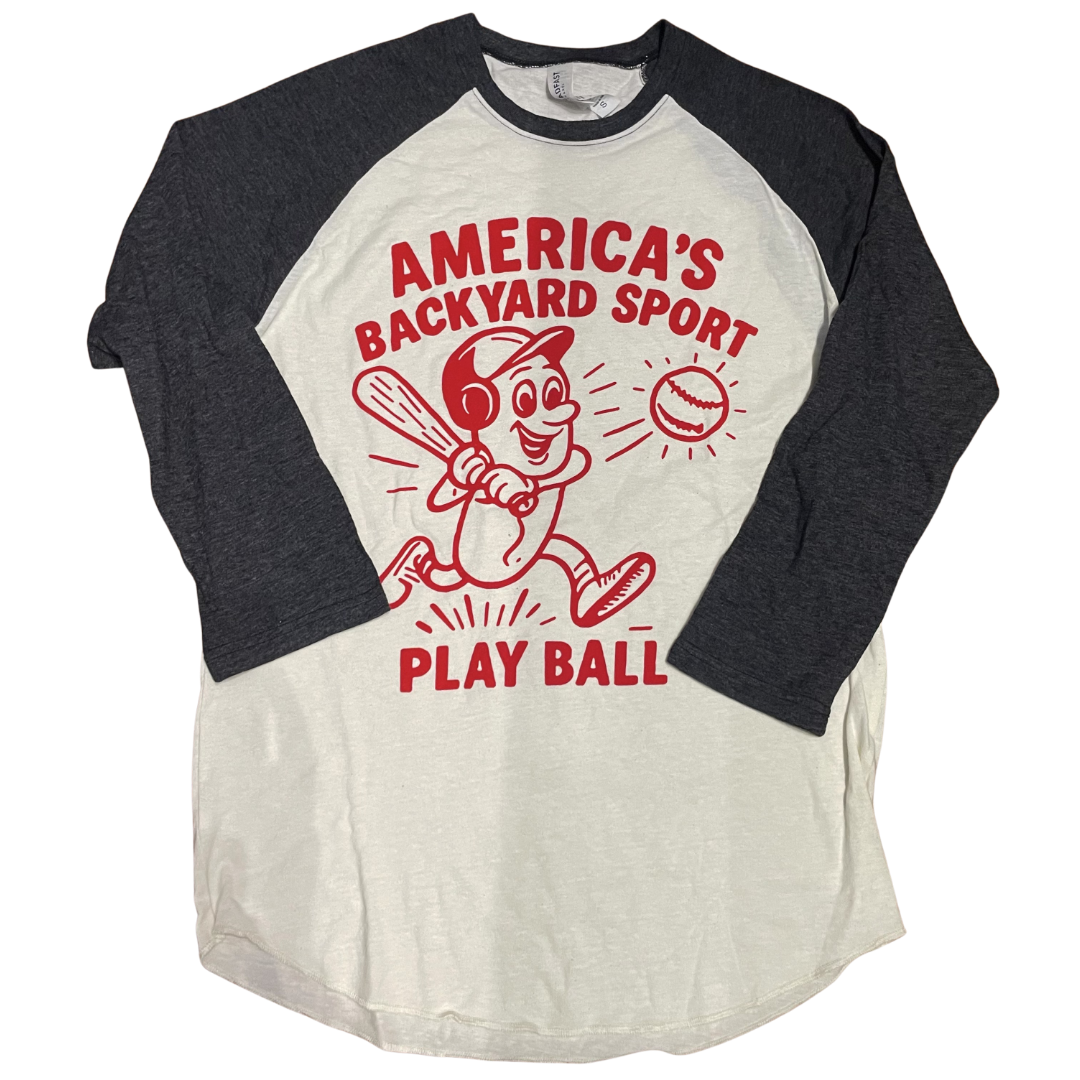 A raglan shirt with a cream torso and graphite sleeves featuring a red design including text that says America's Backyard Sport at the top, a hotdog with a baseball helmet swinging a baseball bat hitting a baseball, and the text Play Ball at the bottom of the design.