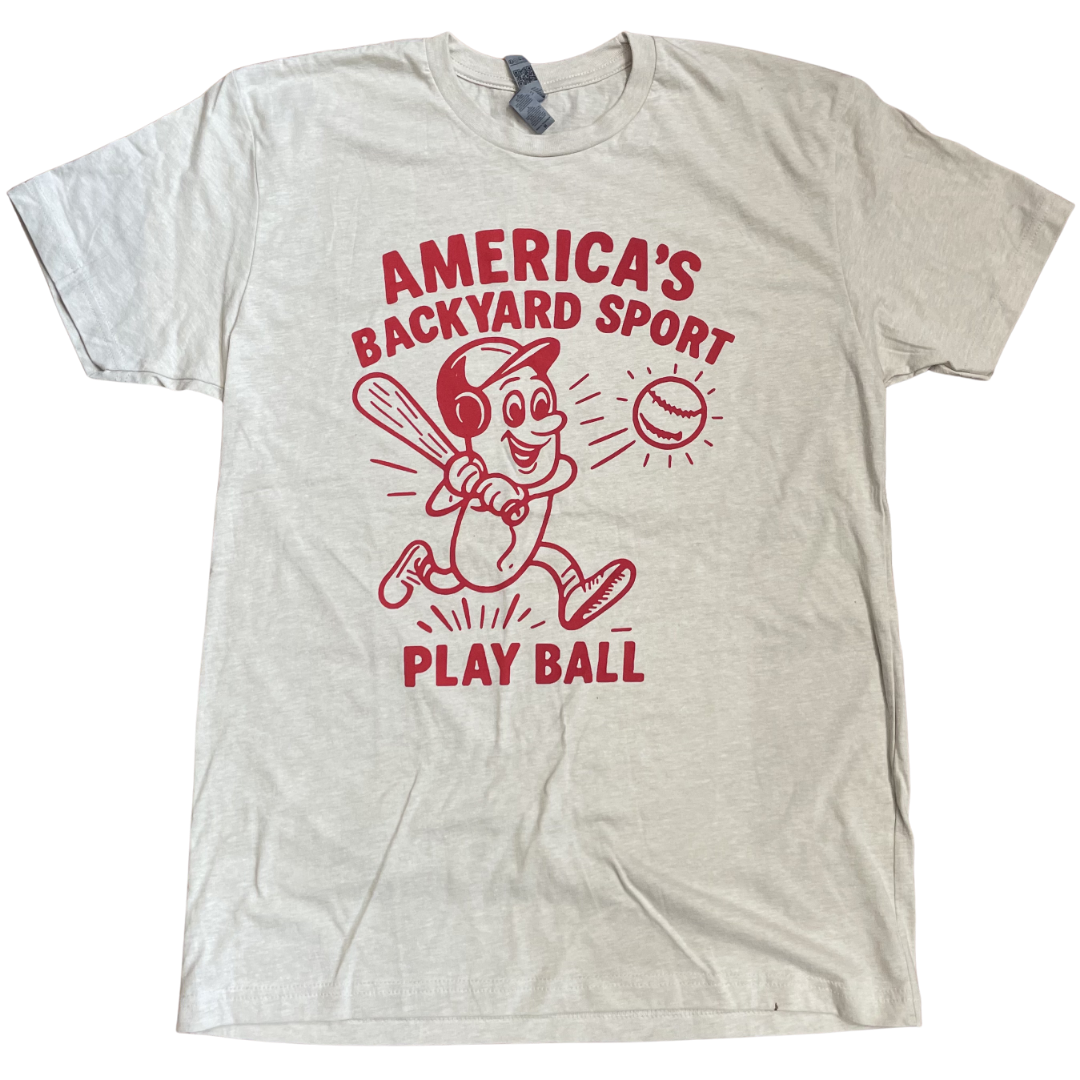 A short sleeve cream tee shirt with a red design including text that says America's Backyard Sport at the top, a hotdog with a baseball helmet swinging a baseball bat hitting a baseball, and the text Play Ball at the bottom of the design.