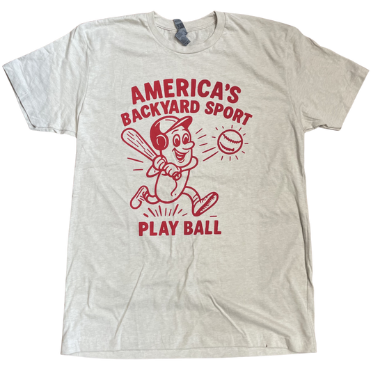 A short sleeve cream tee shirt with a red design including text that says America's Backyard Sport at the top, a hotdog with a baseball helmet swinging a baseball bat hitting a baseball, and the text Play Ball at the bottom of the design.
